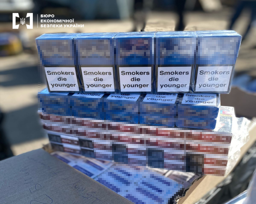 Великим планом декілька блоків сигарет синього та червоного кольору. На пачках попередження про шкоду паління англійською мовою. Назви заблюрено.