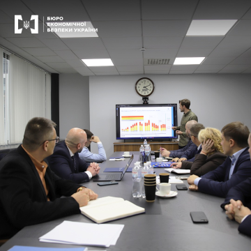 Meeting in a conference room. A group of people in business attire sits at the table, listening to a presentation. A man is showing charts on a large screen.