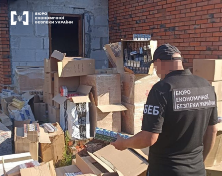 Детектив БЕБ оглядає блок сигарет. На подвір’ї біля гаража довкола нього — велика кількість картонних коробок із сигаретами різних марок.