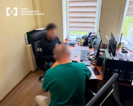 A ESBU detective and a man in a green t-shirt examine papers next to a computer monitor in an office.
