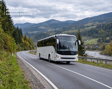 A modern large white bus travels along a two-lane asphalt road that curves against the background of forested mountains and a cloudy sky.