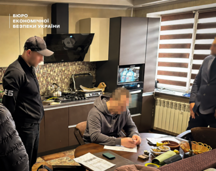 The suspect in the case signs the ESBU search warrant protocol in the kitchen under the supervision of a detective. The man's face is blurred.