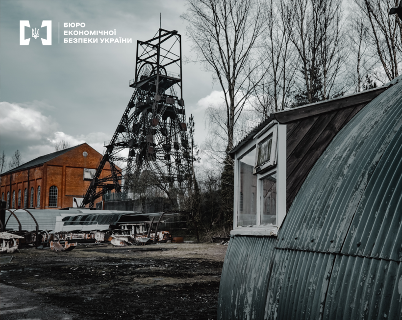 Шахтний копер і промислові будівлі видобувного підприємства серед дерев з оголеними кронами на тлі похмурого неба