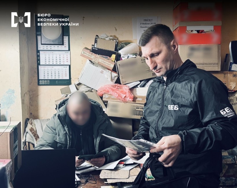 An ESBU detective is standing and examining documents. Nearby, another man, whose face is blurred, is sitting at a desk. The room is cluttered with boxes and papers, and a calendar is visible on the wall.