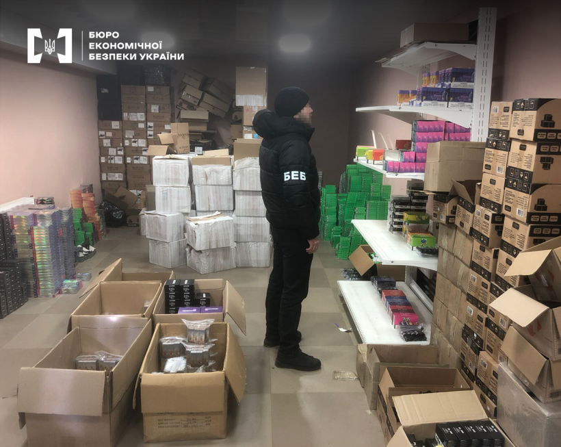 Детектив БЕБ проводить обшук на складі електронних сигарет та рідин. На підлозі стоси картонних коробок різного розміру та стелаж з товаром.