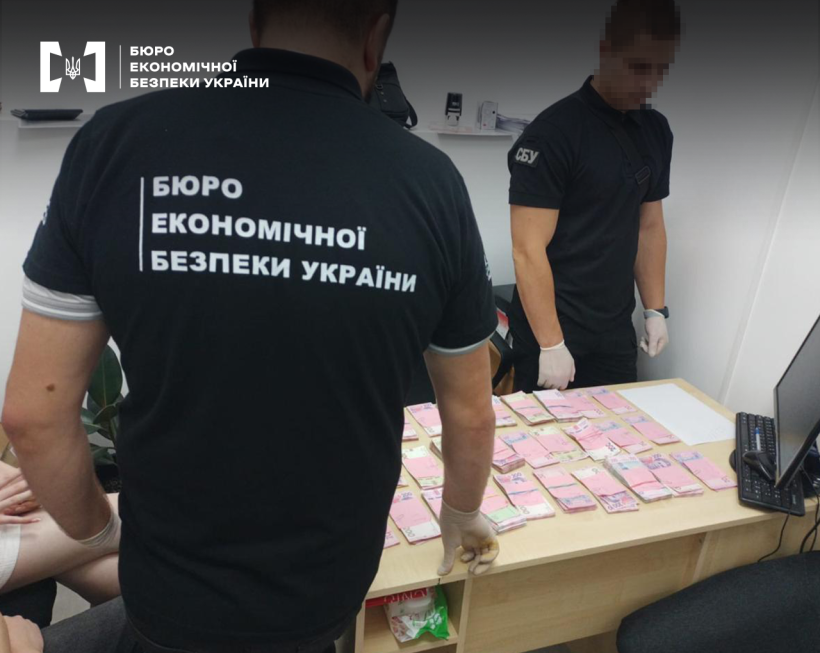 Працівники БЕБ та СБУ проводять обшук в офісному приміщенні. На столі монітор, по усій поверхні столу розкладені пачки 200-гривневих купюр.