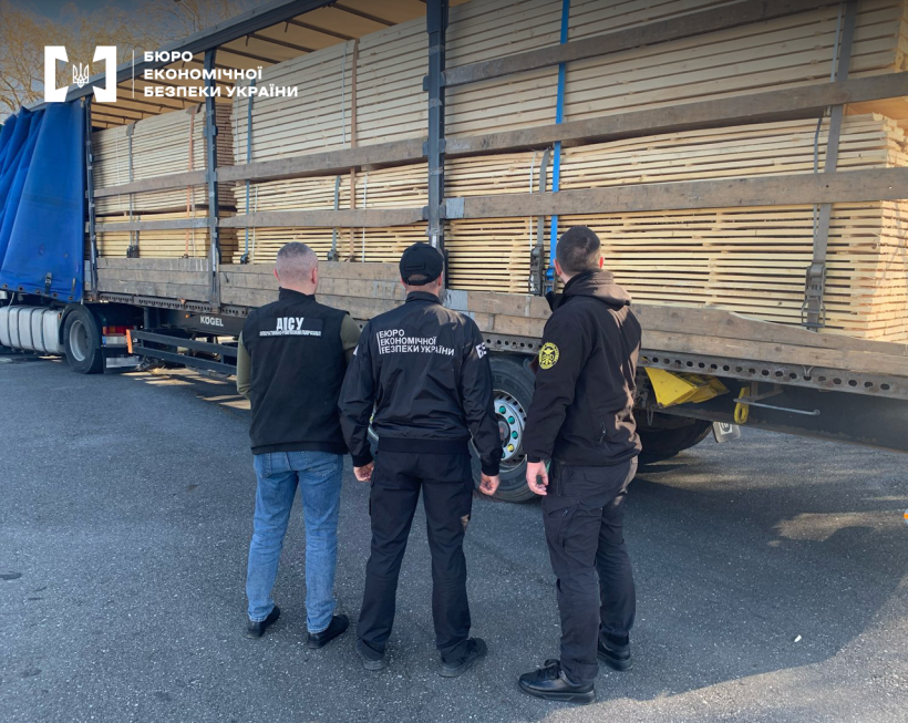 Працівники БЕБ, митної та прикордонної служб оглядають причіп вантажівки з пиломатеріалами, у якого відкрито тент. Дошки щільно складені та закріплені ременями.