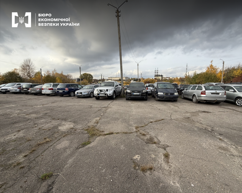 Заасфальтований автомайданчик із рядами припаркованих машин різних марок і типів. На тлі — осінні дерева, стовпи, лінії електропередачі, похмуре небо.