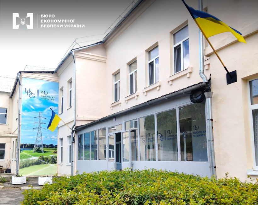 Фасад адміністративної будівлі "Запоріжжяобленерго" із синьо-жовтими прапорами та банером. Перед будівлею зелені насадження