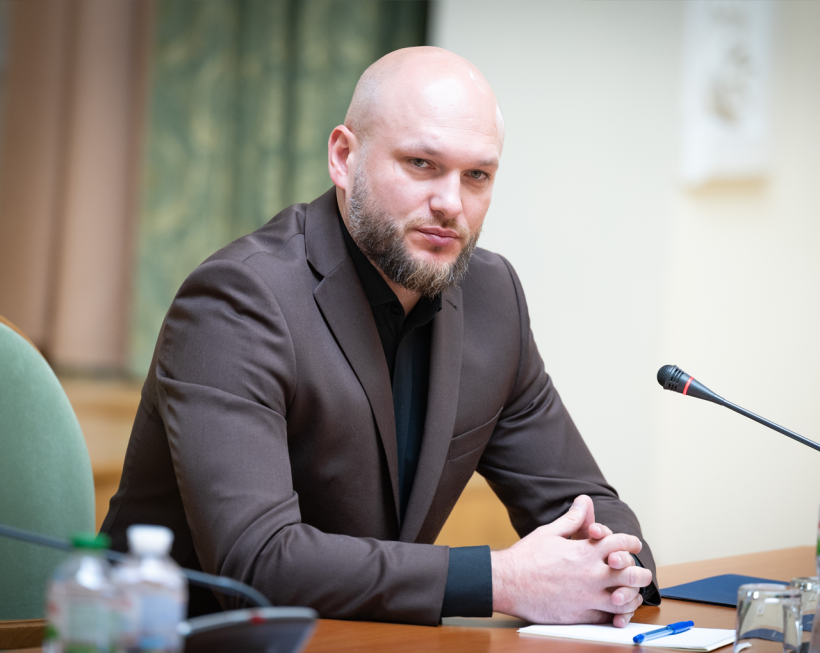 Director of the ESBU, Oleksandr Tsyvinskyi, at a table during a meeting. On the table are a microphone, a notebook, a folder, and bottled water. The background is blurred.
