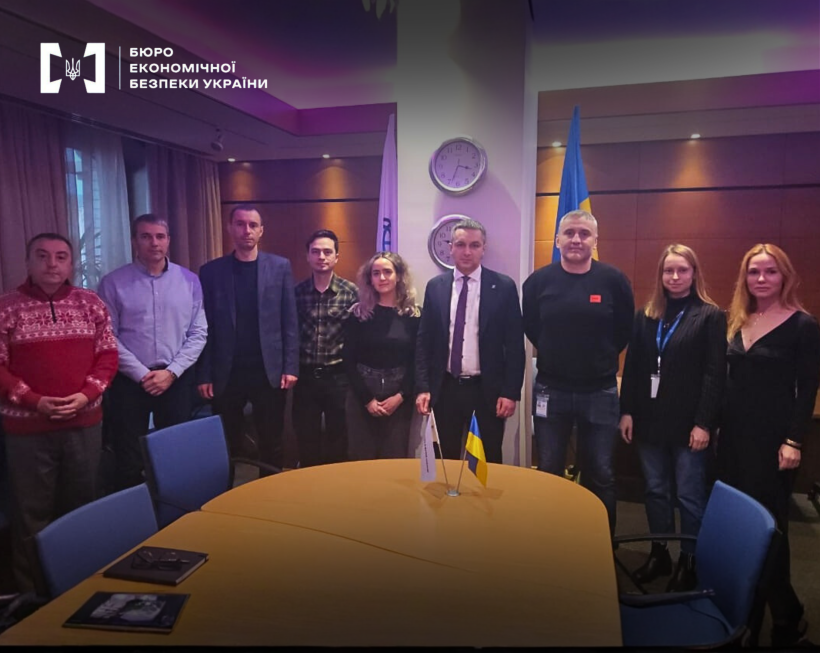 А group of people, including the First Deputy Director of the ESBU, Taras Shcherbai, are standing around a negotiation table with small flags of Ukraine and international partners placed on it