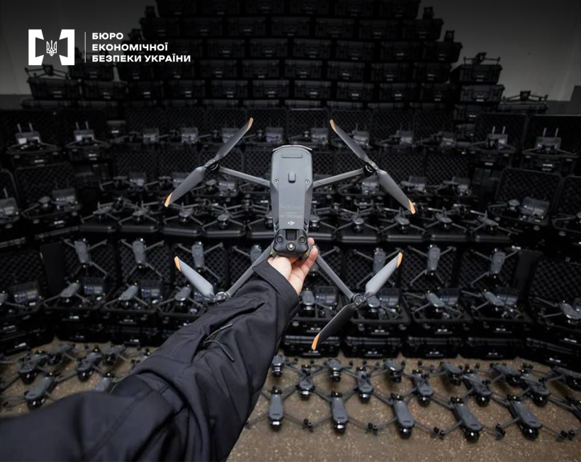 Рука демонструє дрон DJI Mavic на фоні квадрокоптерів у кейсах, складених пірамідою. У нижніх рядах кейси відкриті, кілька апаратів розкладені на підлозі.