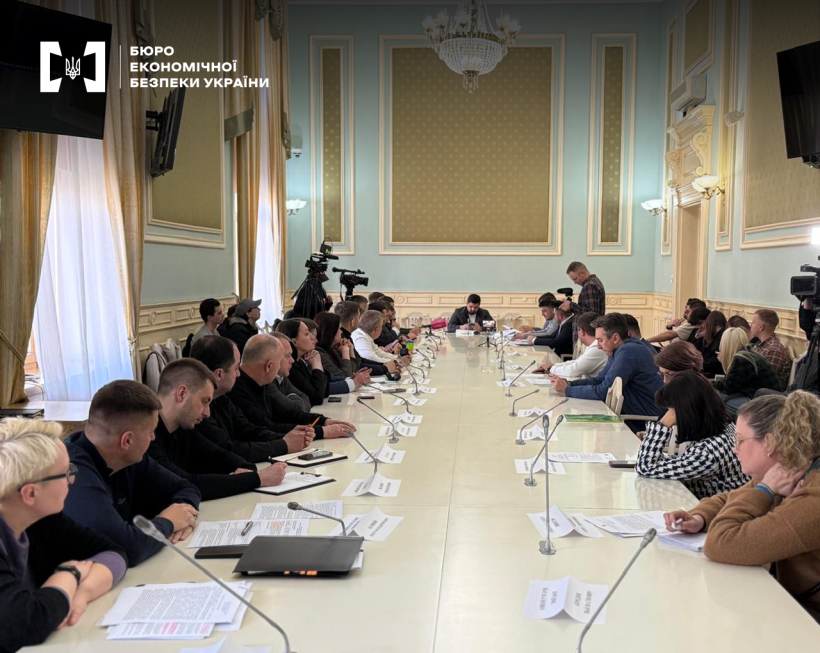 Учасники засідання робочої групи, журналісти й оператори. Просторий зал з високими стелями і класичним інтер'єром.  Довгий стіл, мікрофони, документи