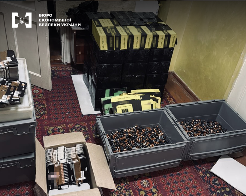 Сірі пластикові контейнери з батарейками, коробки з пакуванням для батарейок, ящики у чорній стреч-плівці на підлозі, вкритій килимом з візерунками.