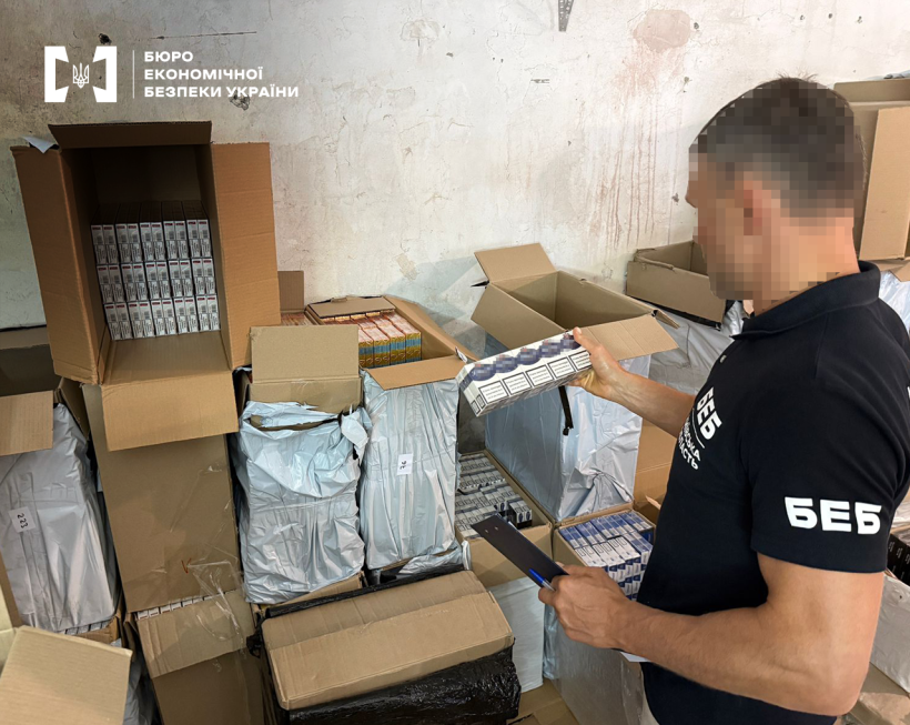 Детектив БЕБ оглядає блок сигарет. Поруч уздовж стіни складені картонні коробки, заповнені сигаретами.