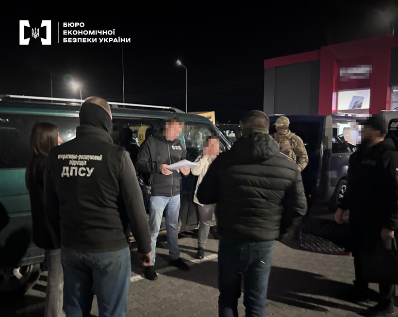 Працівники БЕБ, ДПСУ та СБУ оголошують підозру фігурантці справи. Позаду автомобілі, будівля. Темна пора доби.