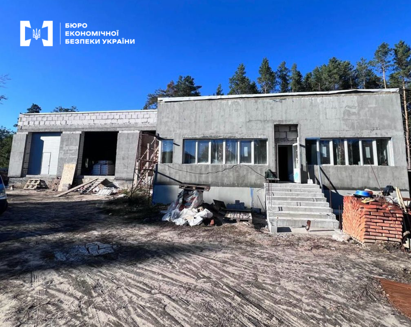 Недобудована будівля пожежного депо, поруч з нею складена цегла, пусті палети, риштування. За будівлею верхівки зелених дерев на фоні синього неба