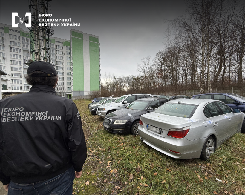 Детектив БЕБ оглядає припарковані просто неба на траві автомобілі. Далі видно житловий комплекс, праворуч – дерева.