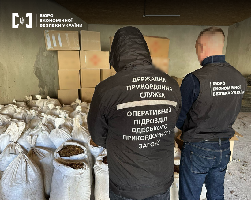 Працівники БЕБ та Держприкордонслужби проводять обшук у складському приміщенні. На підлозі білі мішки з тютюном, стоси картонних коробок.
