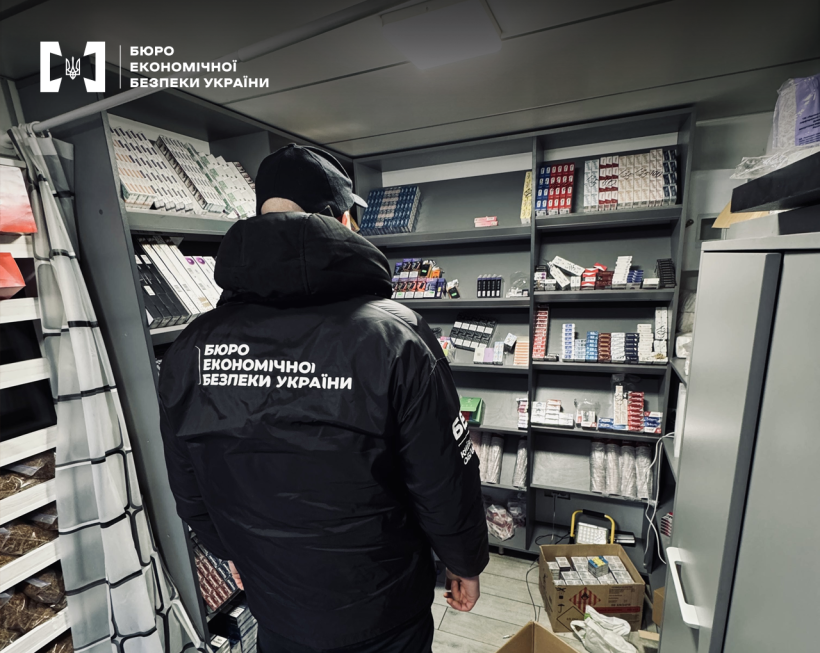 Детектив БЕБ оглядає місце зберігання контрафактного товару. На полицях сигарети, електронки, ємності з рідинами для куріння, розфасований тютюн.