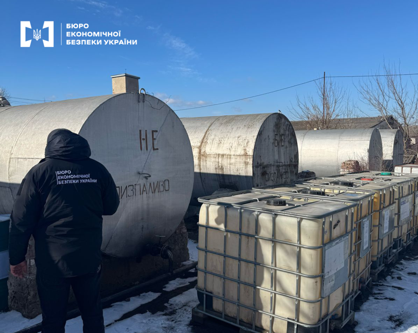 Детектив БЕБ оглядає металеві циліндричні резервуари з дизпаливом, що стоять один за одним. Поруч розміщені резервуари типу «єврокуб» для міні АЗС