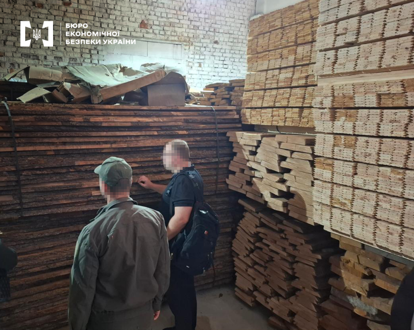 Детектив БЕБ оглядає високі штабелі пиломатеріалів та дощок на складі лісопильного підприємства. Поруч стоїть ще один чоловік.