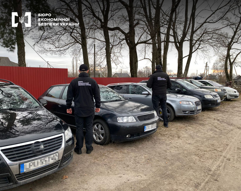Two men in black uniforms stand with their backs to the camera next to several silver and black cars parked on a dirt lot near a red fence