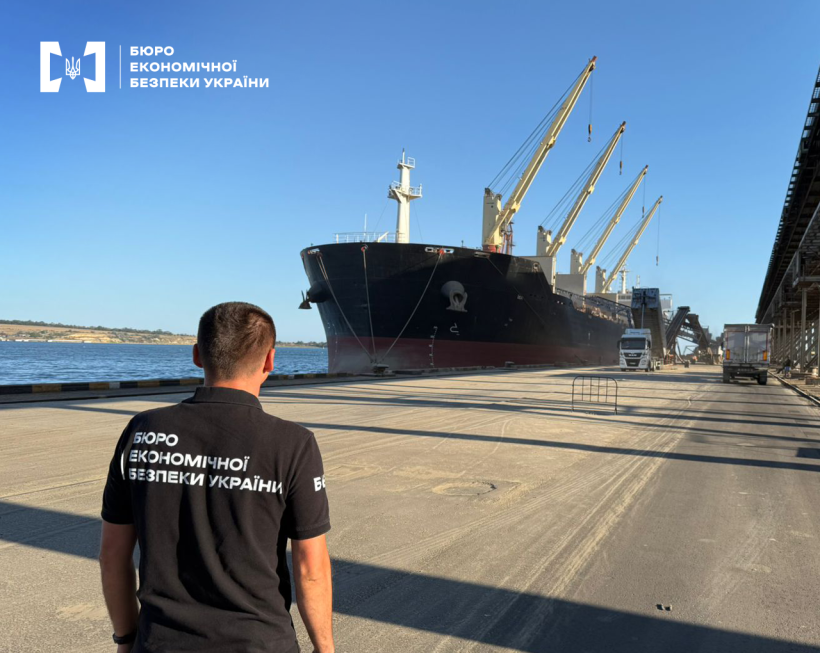 Детектив БЕБ на причалі спостерігає за завантаженням судна. Працюють портові крани, вантажні автомобілі, елеватор. На тлі блакитне небо.