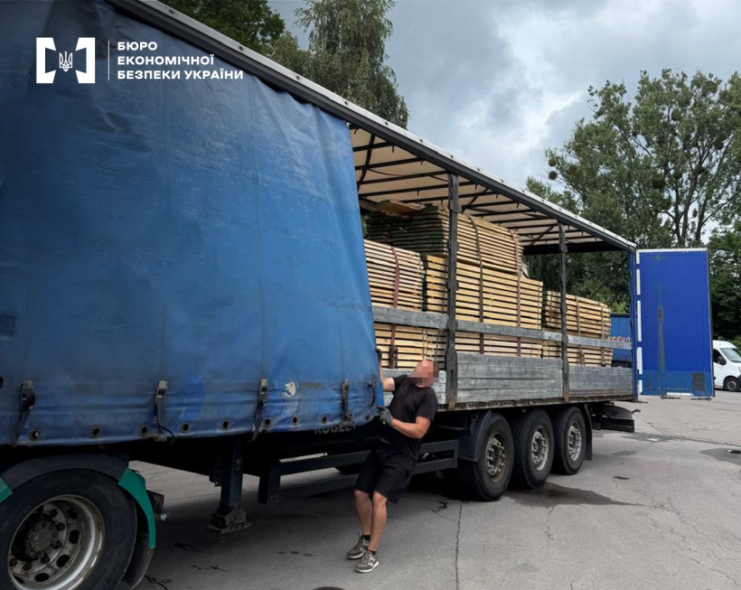 Велика вантажівка з синім тентом та дубовими дошками на тлі дерев та хмурного неба. Водій натягує боковий тент.