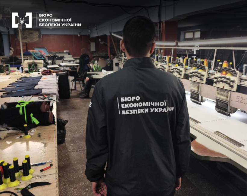 An employee of the ESBU in a black jacket stands with his back to the sewing workshop or production facility with embroidery machines