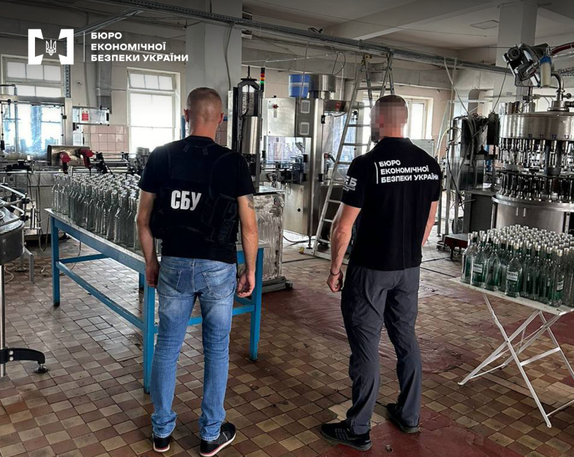 Працівники БЕБ та СБУ оглядають цех з виробництва алкоголю. На столах стоять рядами пусті скляні пляшки, далі — обладнання для розливу.