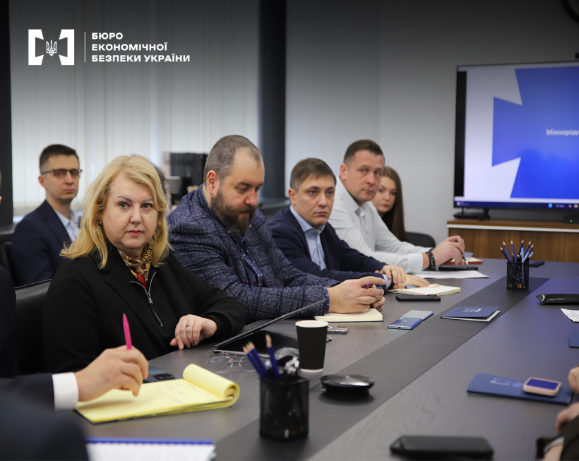 Учасники заходу за столом. На столі документи, записники, папки і блокноти з логотипом БЕБ, олівці й ручки у підставках. На стіні увімкнений екран.