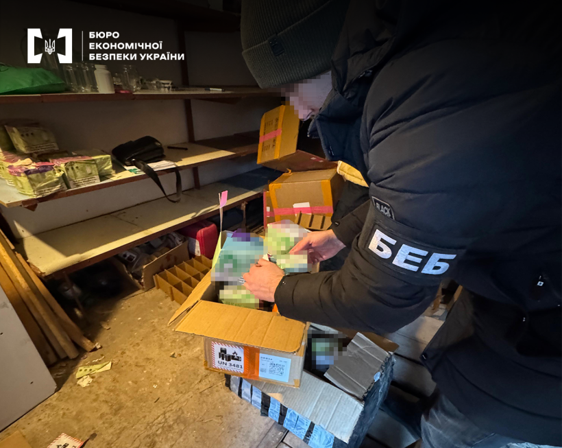 Детектив БЕБ проводить обшук у складському приміщенні, оглядає відкриту картонну коробку. Вміст коробки заблюрено.