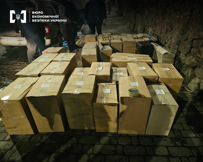 Десятки картонних коробок заклеєні скотчем, що стоять на тротуарі біля кам’яної стіни. Далі поруч з автомобілем кілька чоловіків