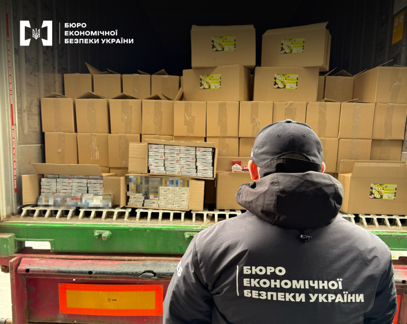 Детектив БЕБ оглядає вантажівку заповнену коробками, частина з них відкрита та містить сигарети. На коробках наклейки із зображенням фруктів ківі.