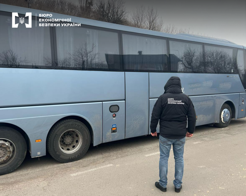 Детектив БЕБ біля рейсового автобуса сполученням «Прага-Краматорськ», що стоїть на дорозі. Верхівки дерев на фоні сірого неба.