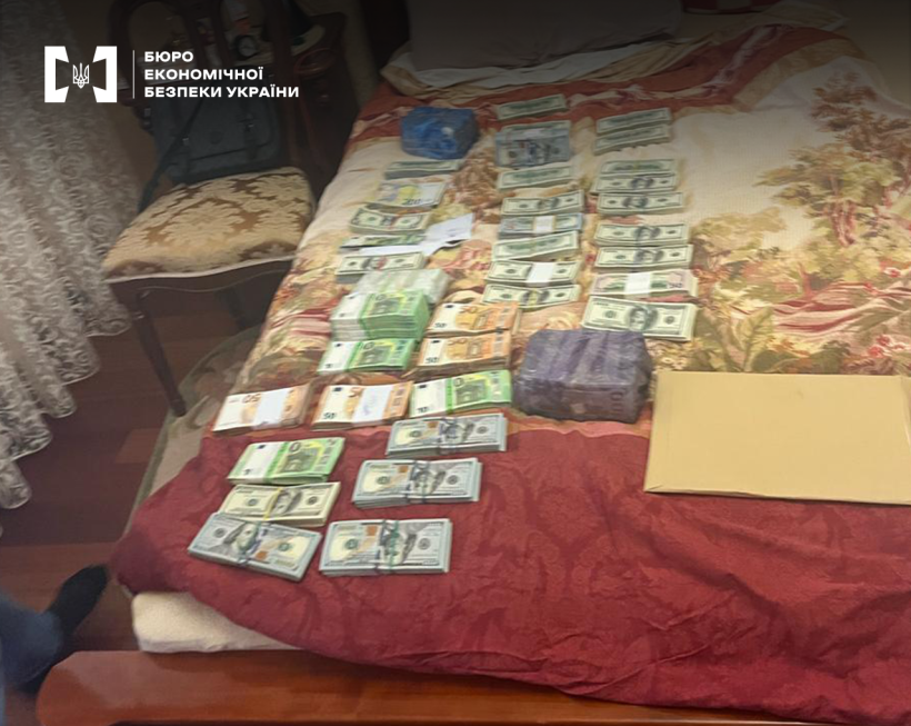 Пачки готівки у доларах і євро різного номіналу розкладені на ліжку під час слідчих дій БЕБ. Поруч дві великі пачки грошей запаковані у плівку, конверт