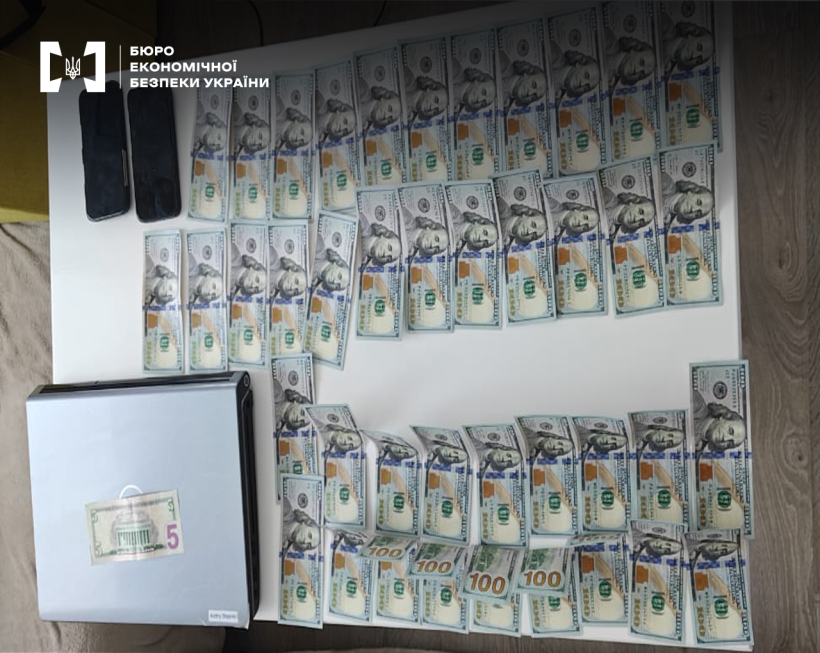 Вигляд зверху. На білій поверхні ноутбук, два смартфони, поруч розкладені фальшиві стодоларові купюри. Одна п’ятидоларова – на ноутбуці
