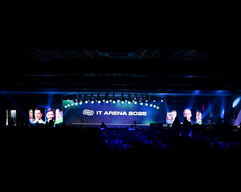 In a dark hall, a brightly lit stage. On both sides of the stage, screens with photos of the speakers; in the centre – the logo and inscription “IT Arena 2025.”