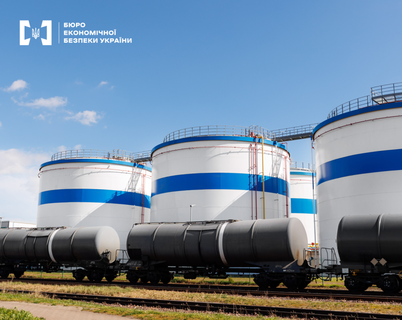 Цистерни на залізничній колії, за ними великі вертикальні резервуари для зберігання нафтопродуктів. Блакитне небо, кілька білих хмар