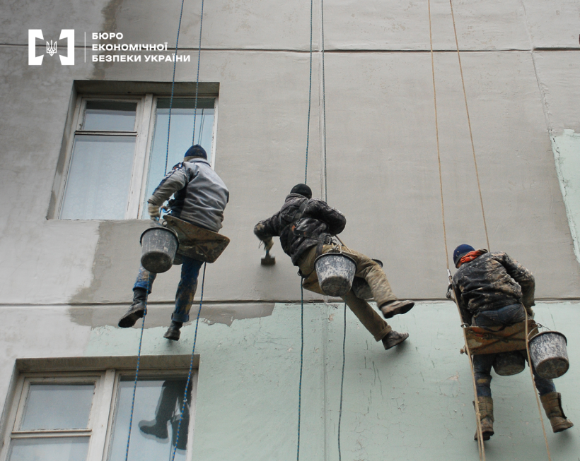 Висотні роботи з ремонту та обслуговування будівель. Троє працівників у альпіністському спорядженні фарбують фасад.