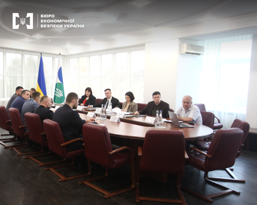 Bright and spacious conference room. Participants seated around an oval table.