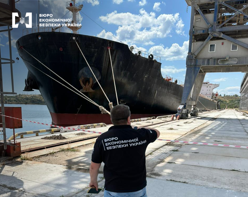 Територія морського порту. Детектив БЕБ на причалі біля вантажного чорного судна та портового крану. На тлі блакитне небо з білими хмарами.