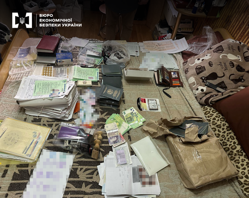 Бланки та печатки різних установ, стоси документів, голографічні елементи, готівка в іноземній валюті розкладені на ліжку під час слідчих дій.