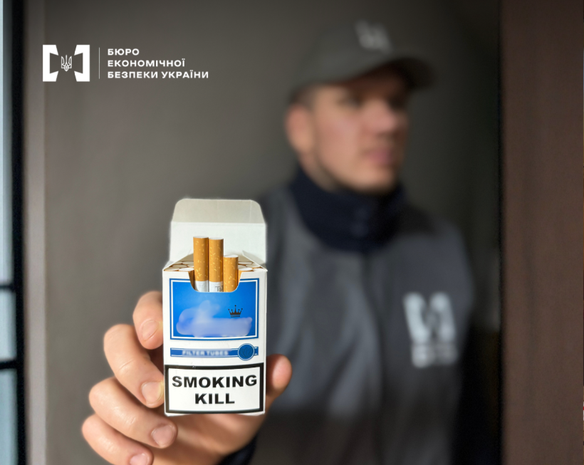 У фокусі відкрита пачка сигарет в синьо-білому пакуванні, яку демонструє Детектив БЕБ.