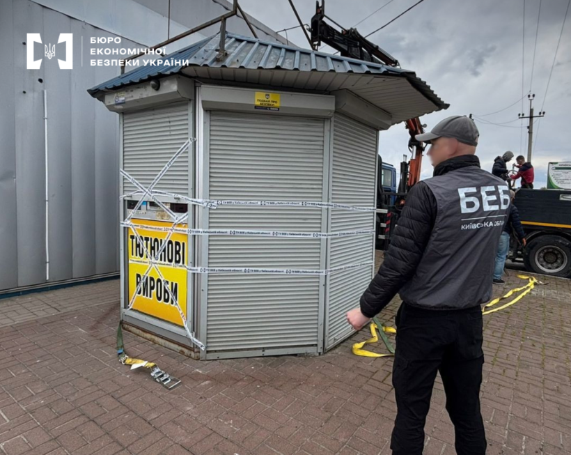 An ESBU detective stands with their back to the camera, observing the dismantling of a gray kiosk. The pavilion features a yellow "Tobacco Products" sign and is wrapped in white tape.