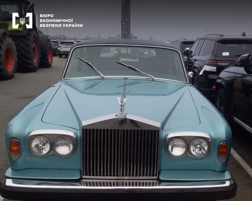 Великим планом передня частина блакитного ретро-автомобіля Rolls-Royce. На задньому плані інші машини та трактор.