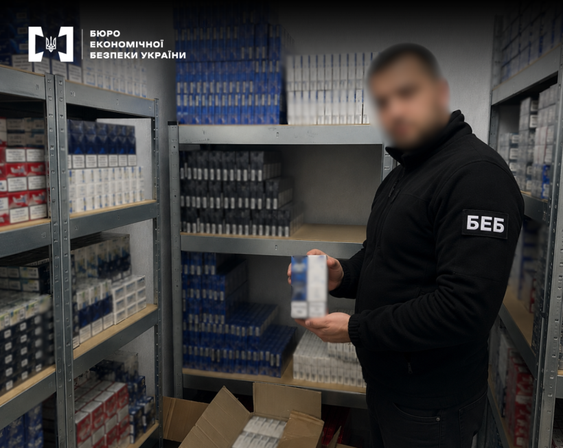Детектив БЕБ демонструє блок вилучених сигарет на тлі стелажів щільно заповнених блоками сигарет різних брендів. Назви заблюрено.