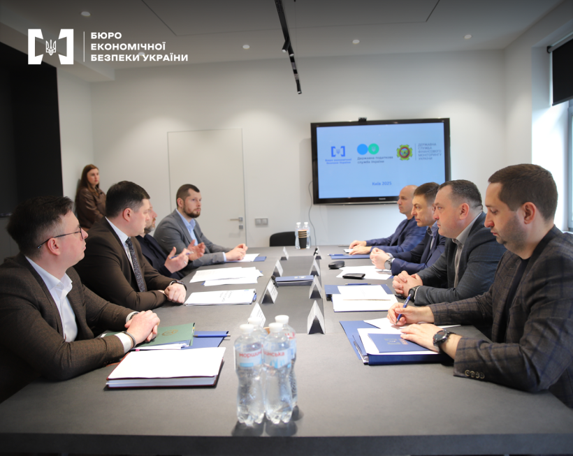 Participants of the event during a discussion in the conference room. On the table are documents, notebooks, water in PET bottles. A screen on the wall shows a presentation