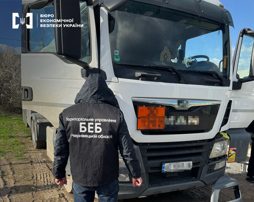 Детектив БЕБ проводить огляд білого великовантажного автомобіля. Номерний знак та марка авто заблюрені.